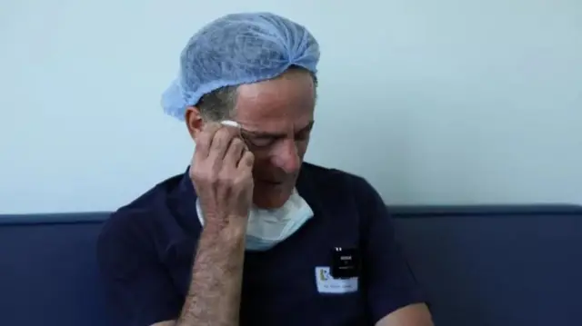 Jaradeh, in scrubs and a hairnet, holds his hand to his temple, looking distressed