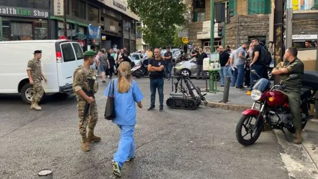 People, including soldiers, stand on a street looking at a robot