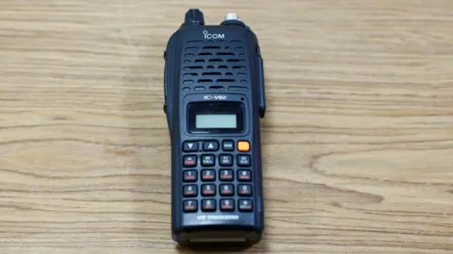 An image of the Icom IC-V82 on a wooden desk. It's a black unit with buttons at the bottom and a speaker/receiver at the top