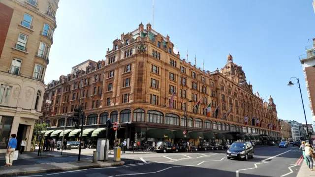 A street-level view of Harrods in London, with black cabs parked outside and the sun shining down