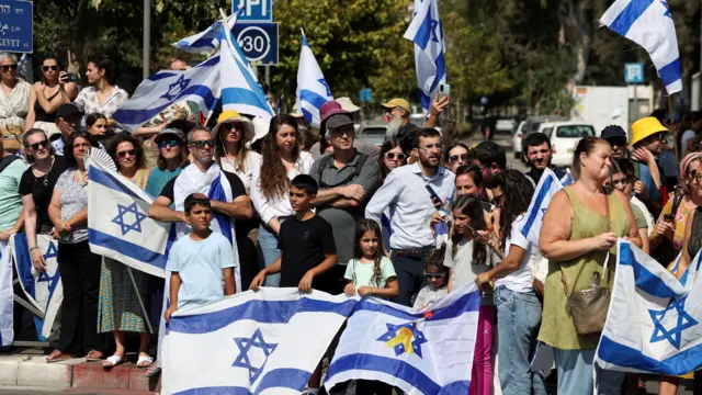 People stand on the side of a street, many holding Israel's flag