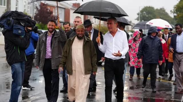 Leaders walk along the road under umbrellas, engaging in conversation