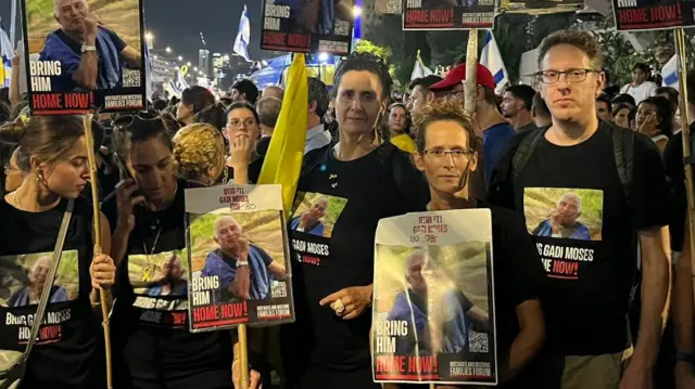 Efrat Machikawa stands in a crowd with other people holding banners and wearing t-shirts with a picture of a man with the text "bring him home now" written on them