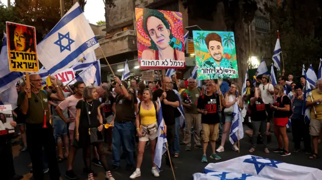 A crowd stands holding banners with paintings of people's faces on them and they are waving Israel's flag