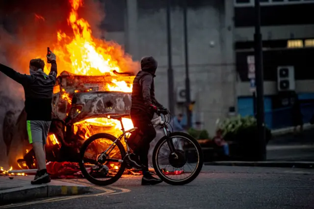 Two people stood in front of a burning car.