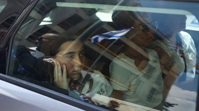 Girl looking sad in back seat of car sat next to two women. A police officer and an Israeli flag can be seen in the reflection of the window.