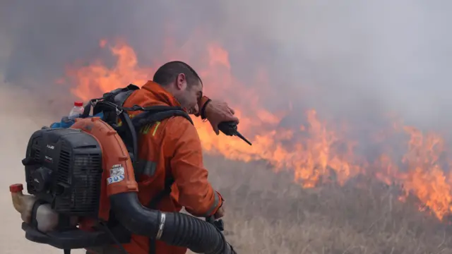 Man wiping his forehead, holding a handheld radio, standing in front of flames