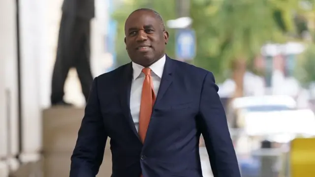 Shot of David Lammy from the waist up, he is wearing a dark blue jacket, white shirt and orange tie.