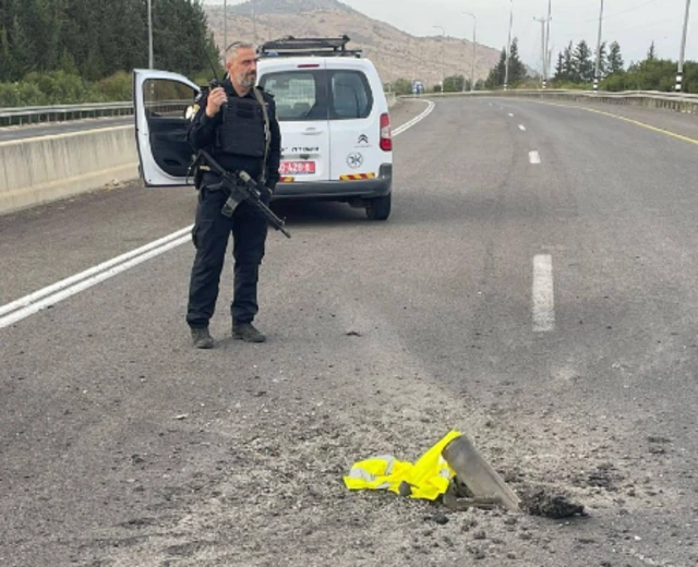 Israeli police officer stands next to some shrapnel from a rocket which has landed in the road. A white van is behind him, and behind him even further is a mountain.