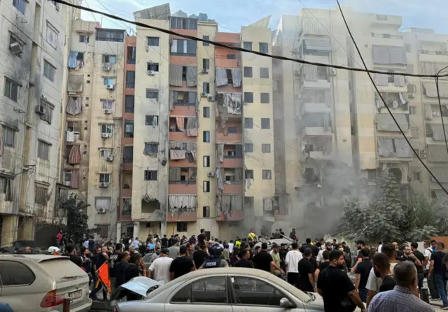 A huge group of people stood in front of a damaged building, smoke can be seen in front of it. Two cars covered in dust are at the front of the picture.