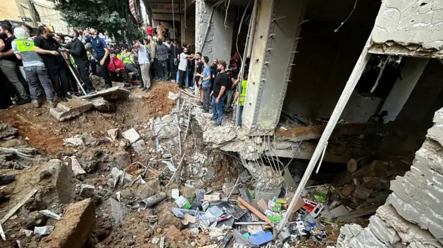 Rubble of a destroyed building, with several items on the floor and a crowd of people in front