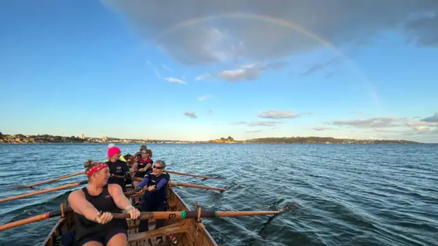 Landscape shot of the ladies' crew rowing where a rainbow can be seen in the background