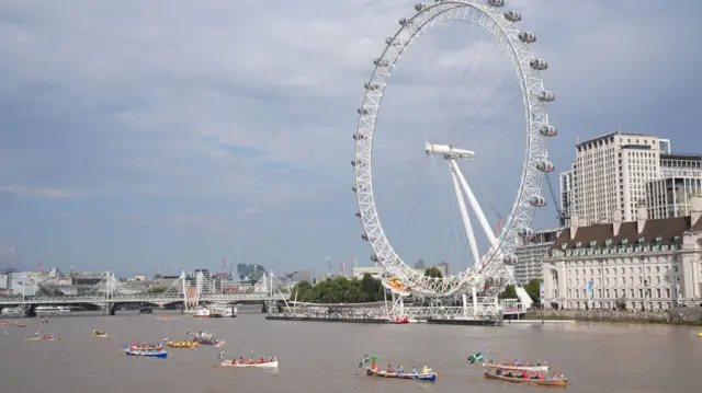 Colourful rowing boats in blue yellow and white boats pass under the London Eye