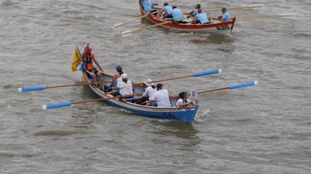 A four-oared rowing boat painted blue and white with blue and white-tipped oars, with four rowers, a cox and passenger