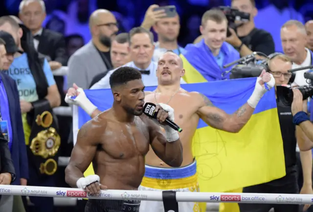 Anthony Joshua speaks into a microphone while Oleksandr Usyk stands in the background holding a Ukraine flag