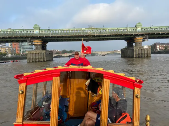 The cabin of the Royal Shallop Jubilant with two men standing at the back. Bridges can be seen in the background