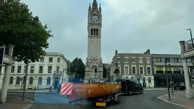 A long boat being towed past a monument on a roundabout on a street