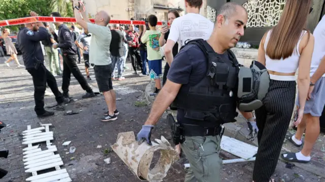 Israeli military specialist carried a piece of rocket while an area is cordoned off with people gathered taking photographs