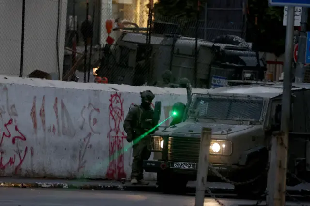 An Israeli soldier stands next to an armoured vehicle shining a green laser