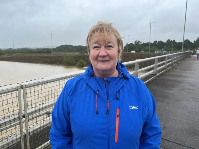 Avril Wright standing on a footbridge overlooking the flooded A421. She is wearing a blue waterproof-type jacket.