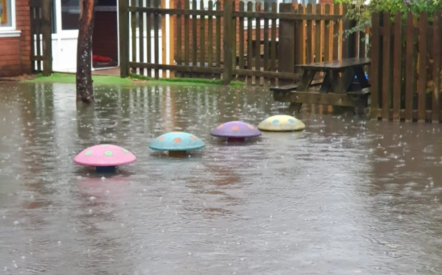 A photo shows the Ardley Hill Academy school playground under water. Four colourful stepping stones in the playground are partially submerged under water as the rain pours. A bench can be seen in the background and a fence.