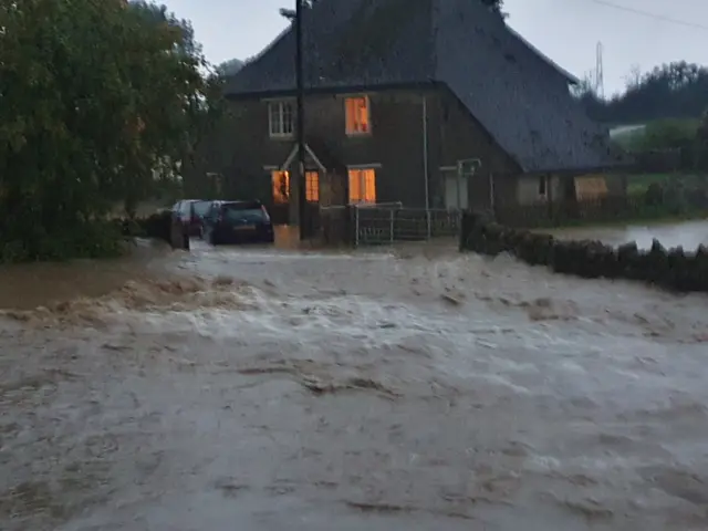 Water shown rushing through Easton Maudit, Northamptonshire, in front of a two-storey detached house which has some of its inside lights on.