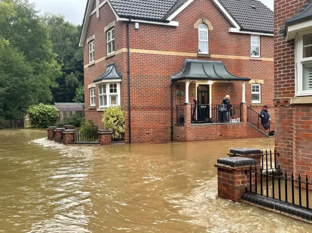 Flood water has submerged a road. Two people can be seen on the steps of a house, overlooking the road, which now resembles a river