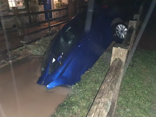 The front of a blue car is submerged in water which fills a hole next to a wooden fence