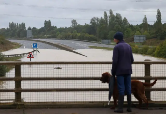 A man walking his dog on a footbridge over the A421. He is looking down at the flooded road where part of a car is peeking above the flood water.