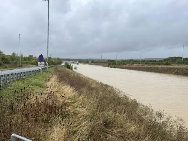 A photos shows the flooded A421 road from a side angle. Vehicles can be seen in the distance avoiding the flood water.