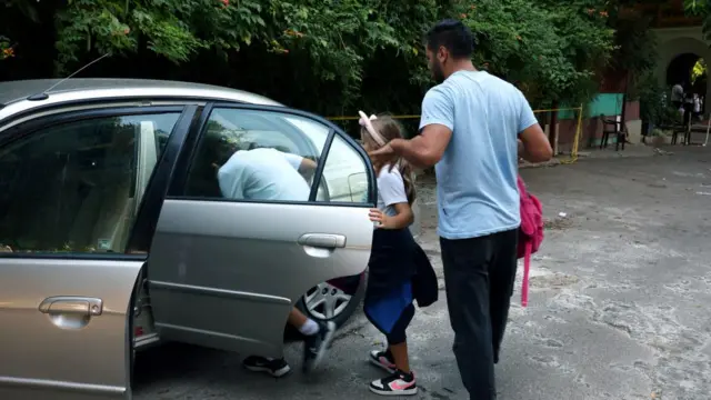 A man opens the back door of a silver car for two school-age children