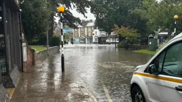 A photo taken from the side of a road in Buckinghamshire. It shows the road partially flooded while a van sits at the side of the road. It is raining and the flood water has in parts broken onto pavements.