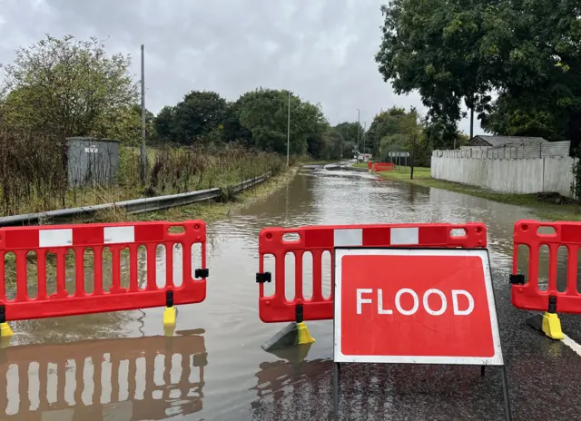 A photo shows a flooded road in Dunstable that has been cordoned off with signs warning of floods. The flood water stretches about 100 metres.