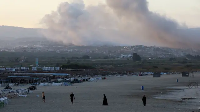 Smoke drifting over a town with people walking along beach in foreground