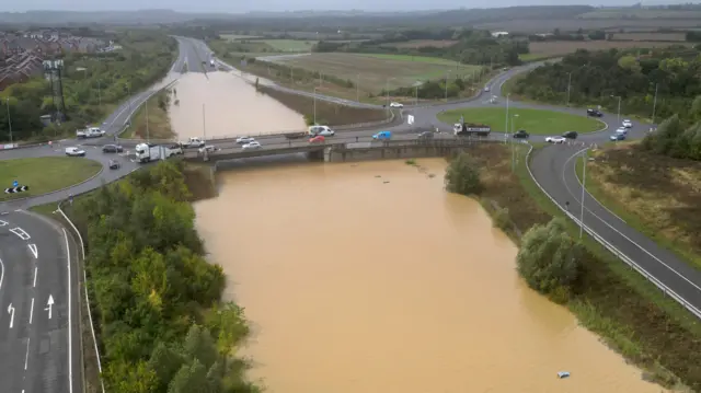 Huge flooding submerges the A421, with a car boot poking out of the muddy floodwater in the bottom right of the photo