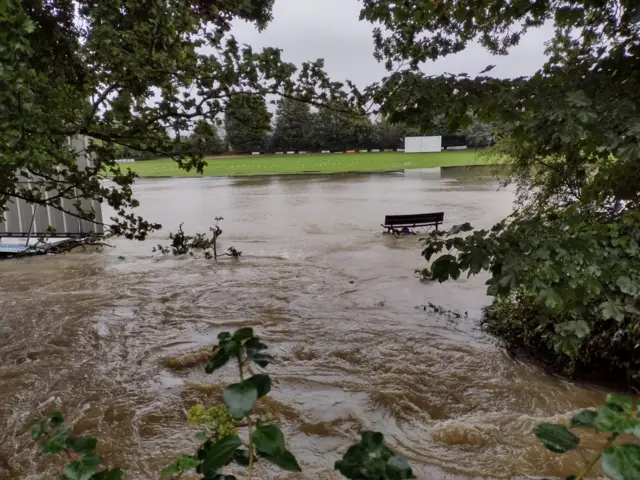 The photos show the River Flit which has burst its banks. Water can be seen flooded around the area and onto a nearby cricket pitch. A bench has been submerged in the water.