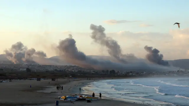 People walkign along a beach with smoke in the background.