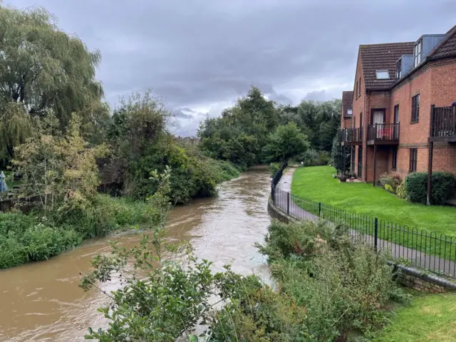 The river is rising, you can see some bricks below a path at the bank of the river, next to some houses.