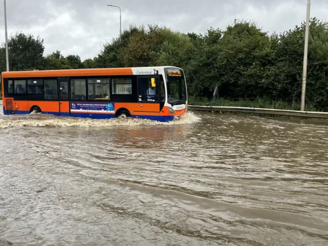 An orange and white bus struggling to get through part of a flooded road.