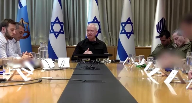 Yoav Gallant addressing a meeting in Tel Aviv with officials sitting in front of him and Israeli flags behind him