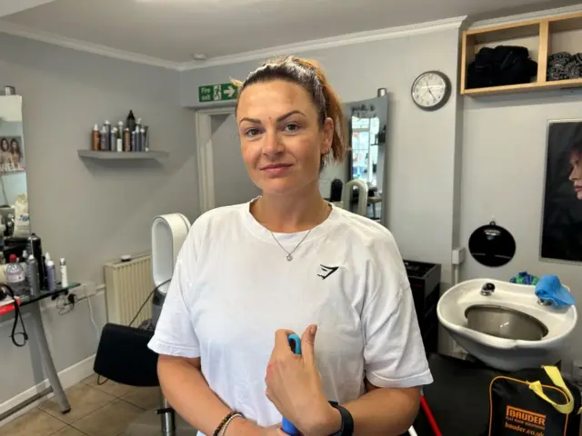 Kirsty Brewer is standing in a hair salon. She is wearing a white T-shirt and her dark hair is pulled back into a ponytail. She is holding onto a broom.