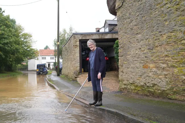 Carol Maher is pictured dipping a mop into flood water outside her house Grendon. She is looking toward the camera while wearing Wellington boots and warm clothing.