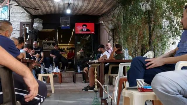 Men sit on plastic chairs next to lines of wooden tables in a cafe in Beirut. They are drinking bottles of water and some of them are looking to watch a television screen in the corner with the face of Hezbollah's leader Nasrallah.