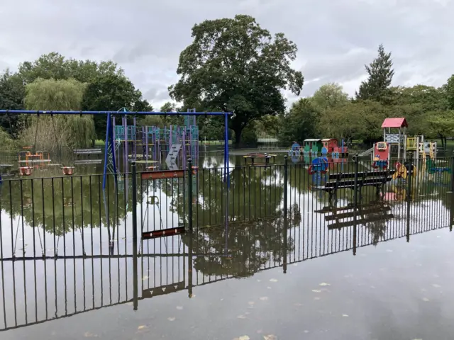 The picture shows Luton's Wardown Park and children's play area submerged in water. Playing equipment can be seen which is behind a fence.