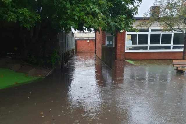 A photo taken from outside Ardley Hill Academy school in Dunstable, Bedfordshire. It shows the exterior of a red brick school building that has been surrounded by water. Rain drops can be seen hitting the water.