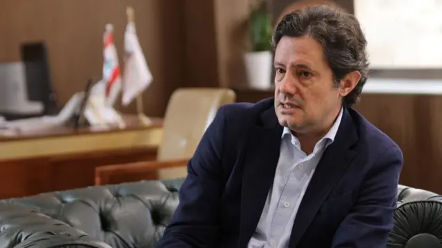 A man in a suit and blue shirt sits on a leather sofa and looks at the camera. A desk with a Lebanon flag on it is behind him