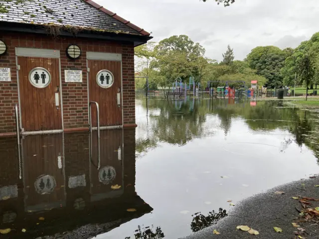 A wider photo of Wardown Park in Luton shows the public toilets also surrounded by water. The playground can be seen in the distance under water.