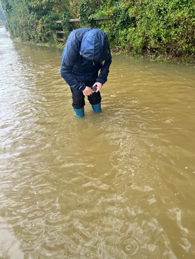 A person wearing a rain jacket looks down to take a picture of the floodwater they are standing in. The water reaches nearly to the top of a pair of wellington boots they are wearing.