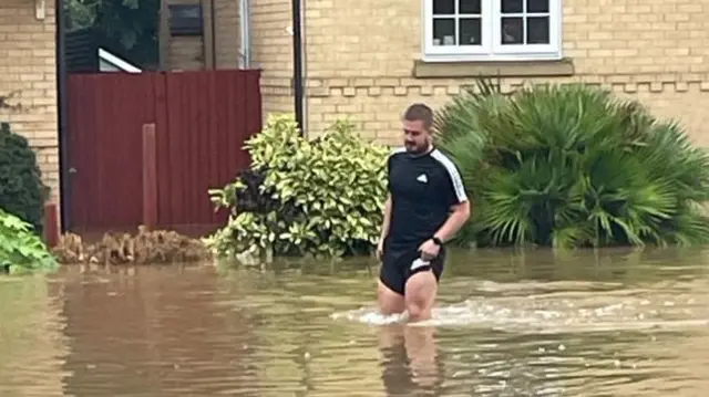 A man wearing a black T-shirt hoists up his black shorts as he walks through flood water. The water can be seen gushing up to above his knee level