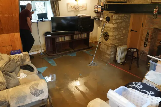 A photo taken from inside Tim Maher's home that was flooded in Northamptonshire. He is pictured on the phone looking out of the window while flood water lies around him. A mop can be seen sat against the wall while boxes of items sit on the sofas.